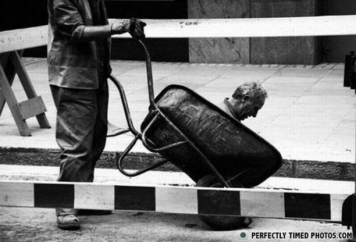 Man sitting inside a wheelbarrow being pushed by another person in a construction area, a perfectly timed photo.