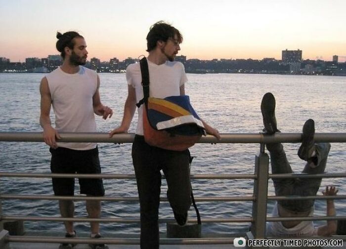 Two men near a waterfront railing with a third person caught in a perfectly timed photo falling upside down over the edge at sunset.