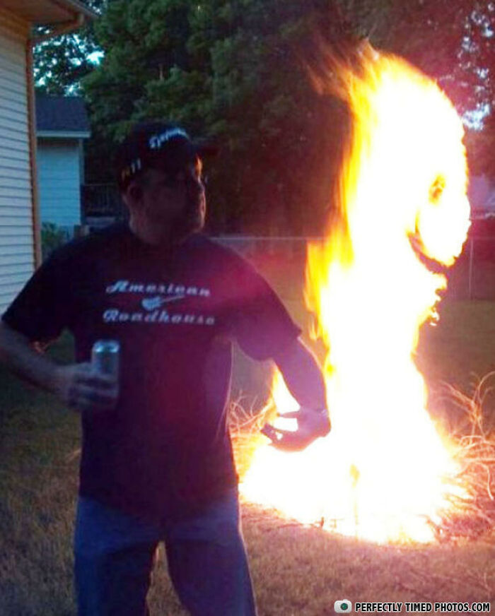 Man holding a drink captured in a perfectly timed photo with a fire shaped like a smiling face in the background outdoors.