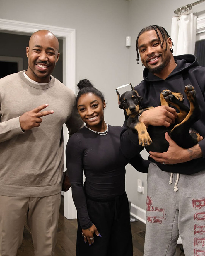 Simone Biles standing between two men indoors, while one man holds a dog with cropped ears in his arms. Simone Biles standing between two men indoors, while one man holds a dog with cropped ears in his arms.