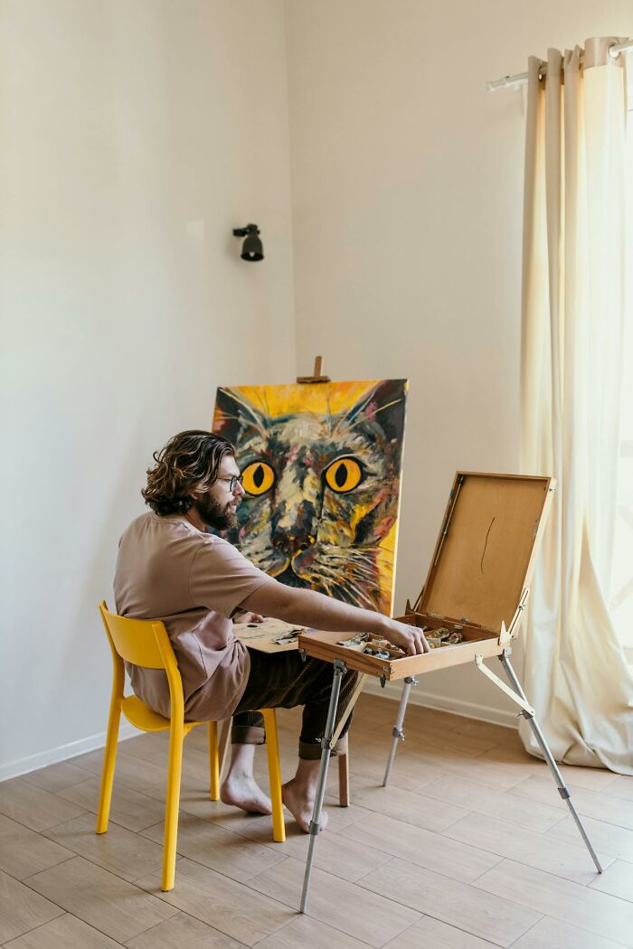 Man sitting on yellow chair painting a large colorful cat portrait, demonstrating creativity and artistic entitlement.