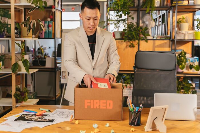 Man packing belongings in a box labeled fired, illustrating sneaky acts of revenge in a modern office setting.