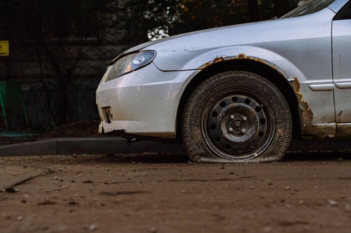 White car with a flat tire and rusted wheel well parked on a dirt road showing a sneaky act of revenge.