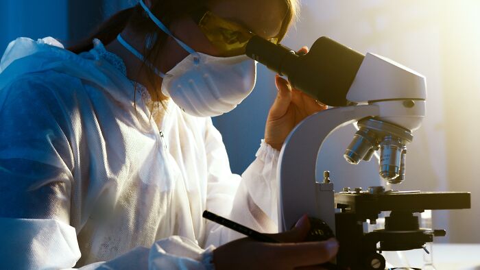 Scientist wearing protective gear and mask, examining samples under a microscope in a lab focused on unresolved mysteries.