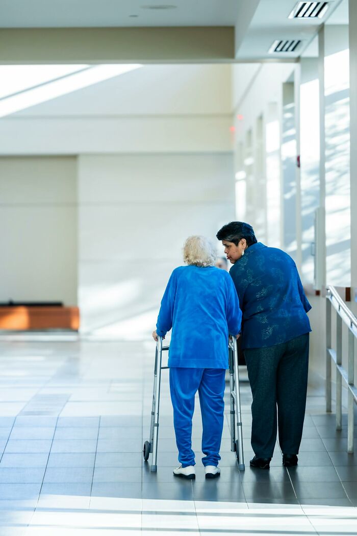 Elderly woman using a walker with assistance in a bright hallway, illustrating disturbing facts people learned against their will.