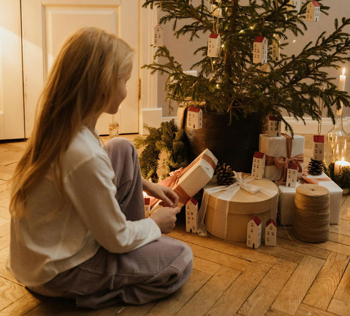 Young girl sitting by Christmas tree holding a gift in a cozy room with wrapped presents and festive decorations.