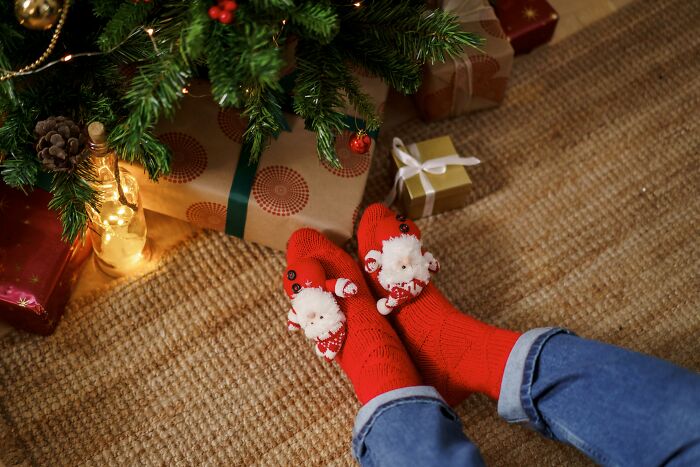 Person wearing red Christmas socks with Santa decorations near wrapped gifts under a decorated Christmas tree.