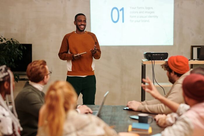 Man in brown sweater delivering a clever and funny presentation to a diverse group in a modern meeting room.