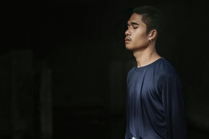 Young man in a dark blue shirt standing with eyes closed against a black background in a quiet moment at the airport.