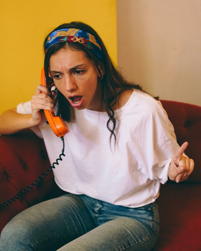 Young woman showing pure entitlement, angrily talking on an orange phone while sitting on a red couch indoors.