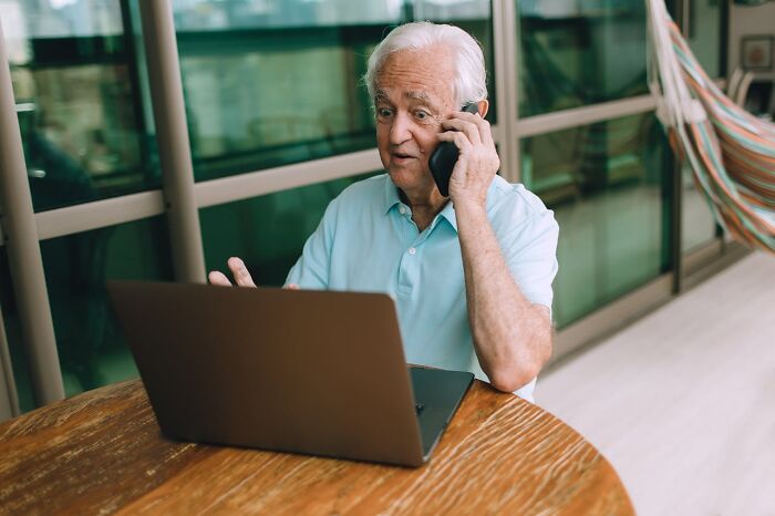 Elderly man talking on phone while using laptop at a wooden table, illustrating sneaky acts of revenge concept.
