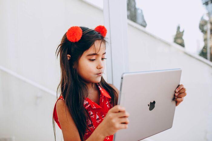 Young girl with red hair accessories and dress, looking intently at a tablet, showing pure entitlement expression.