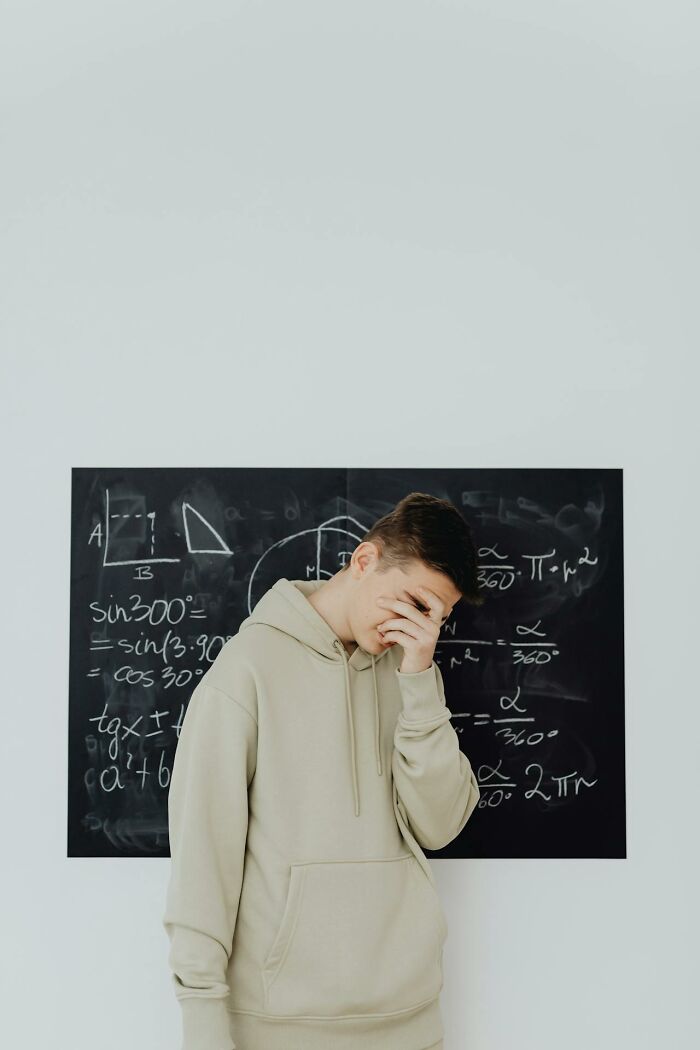 Teen student in beige hoodie covering face in front of math equations on blackboard, showing funny and sassy moment.