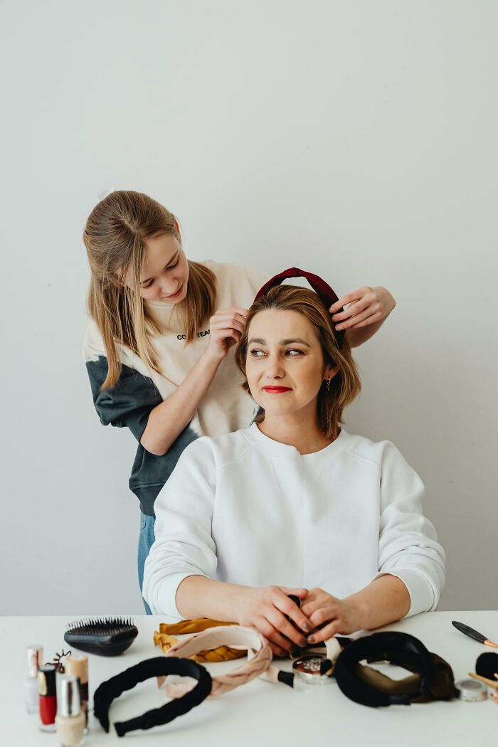 A daughter helping her mother try on modern parenting trends with various headbands and makeup on the table.