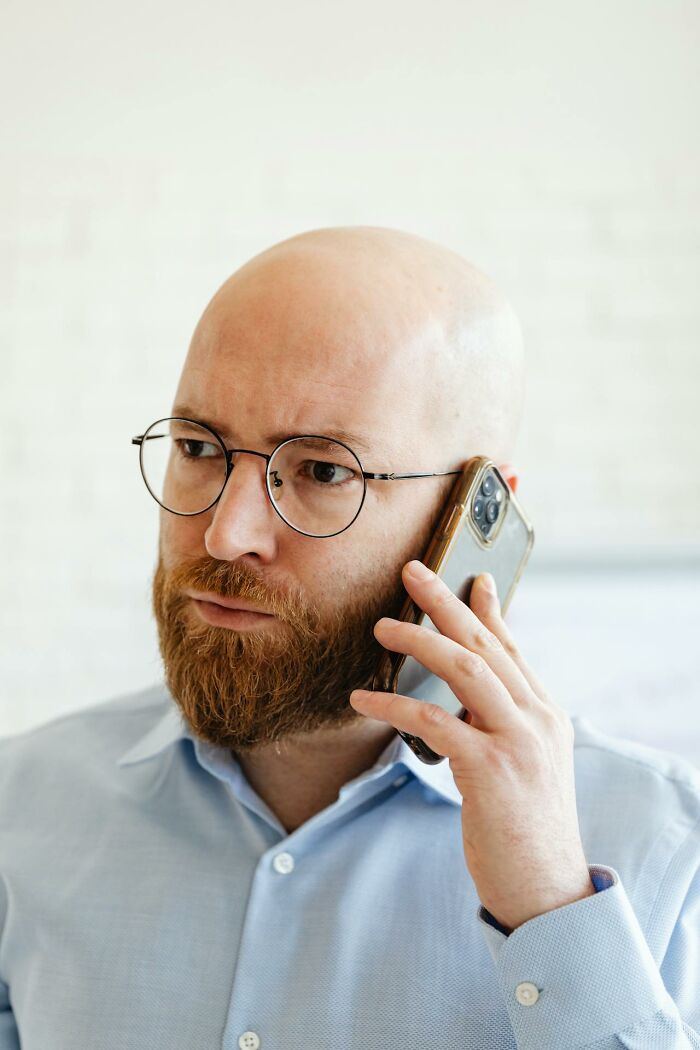 Bald man with glasses and beard in blue shirt making a phone call, representing brilliantly sneaky acts of revenge.