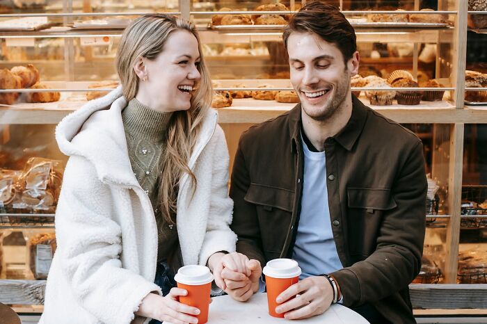 Couple laughing and holding hands at a bakery, sharing funny, clever, and sassy moments over coffee.