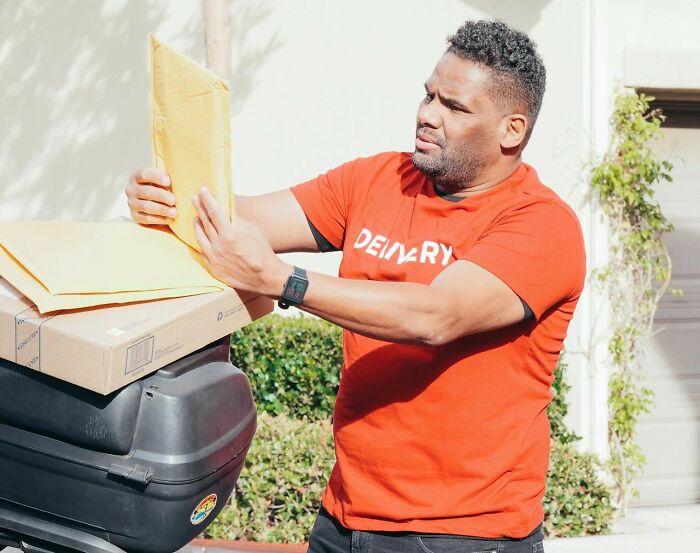 Postal worker in a red shirt sorting and delivering packages outdoors during a sunny day.