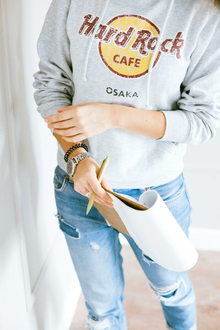 Person in casual hoodie and ripped jeans holding clipboard and pen, capturing funny clever sassy moments on paper.