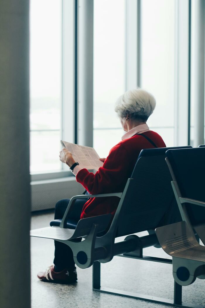 Elderly woman in red sweater sitting in airport waiting area reading a newspaper, typical airport stories scene.