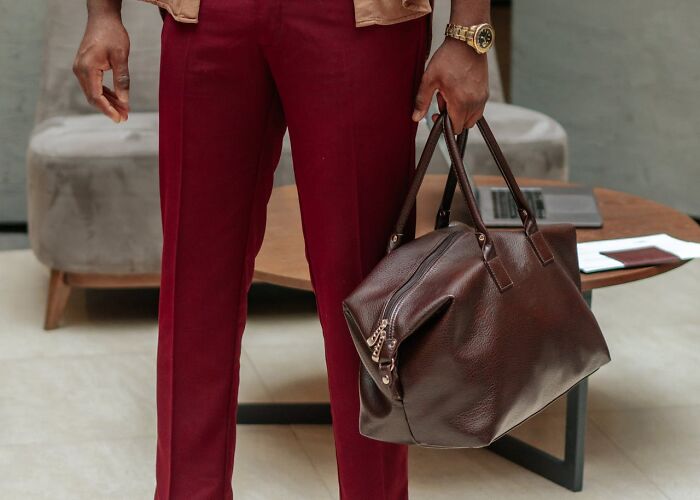 Man holding large brown leather bag, wearing red pants and gold watch, standing indoors near table and chair, crazy airport stories.