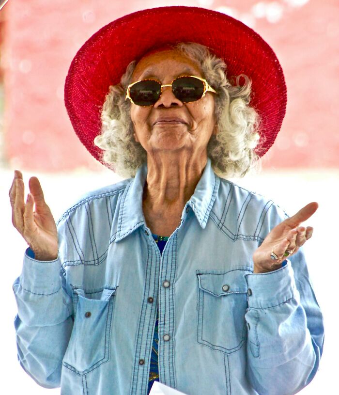 Elderly woman in sunglasses and red hat gesturing with hands, reflecting on crazy airport stories and travel experiences.