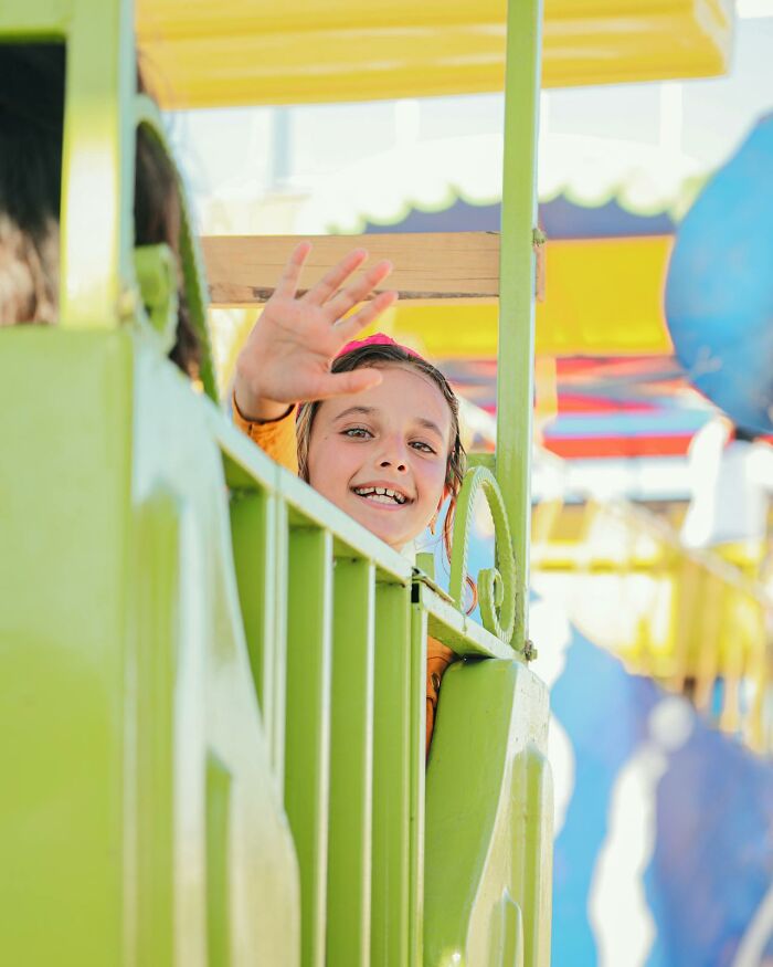 Smiling young girl waving from a colorful amusement ride with bright yellow and green details in an outdoor setting.