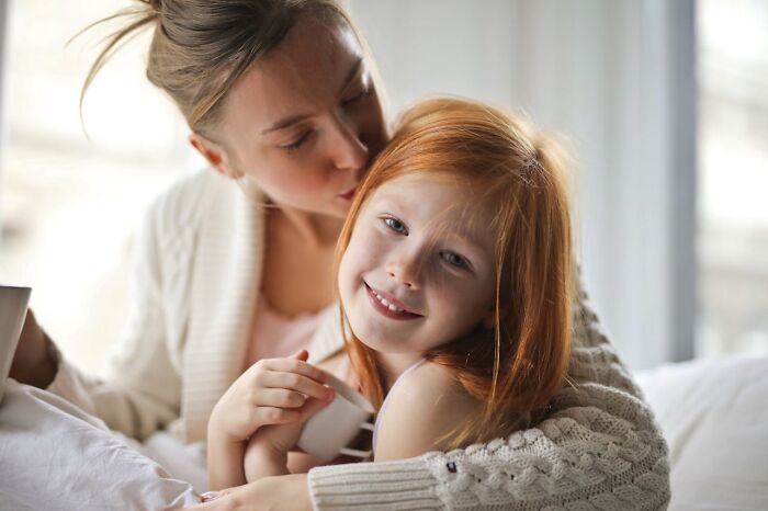 Mother showing affection to her smiling red-haired daughter, illustrating modern parenting trends in a cozy home setting.