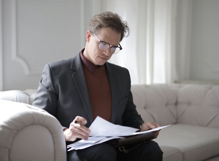 Man in glasses and suit sitting on couch reviewing disturbing facts in documents with focused expression
