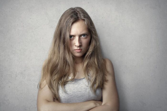 Young woman with long hair crossing arms and displaying pure entitlement with an intense, angry expression against neutral background.