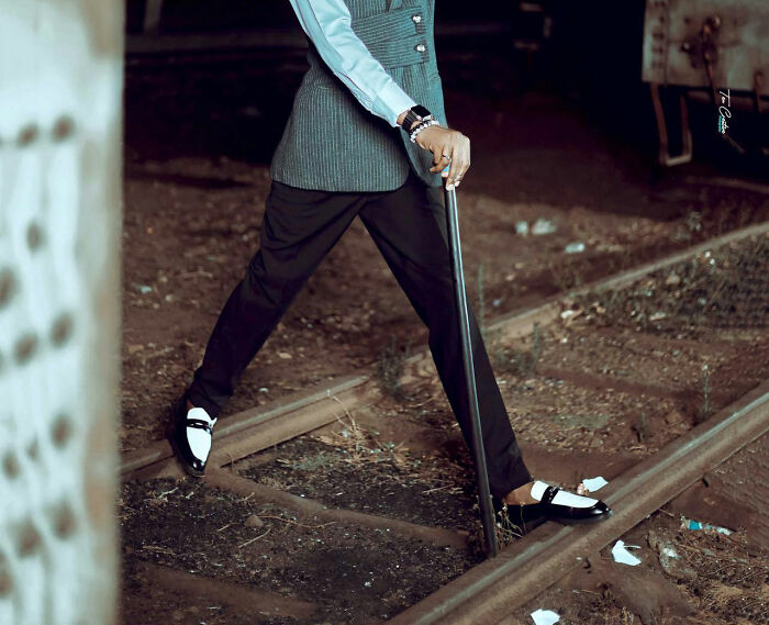 Person dressed formally holding a cane, standing on old railway tracks among unusual personal belongings left behind.