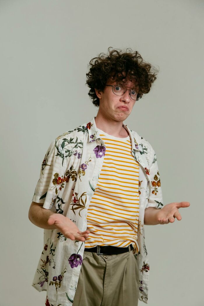 Young man with curly hair and glasses wearing floral shirt and striped tee, shrugging with a confused expression, airport stories theme.
