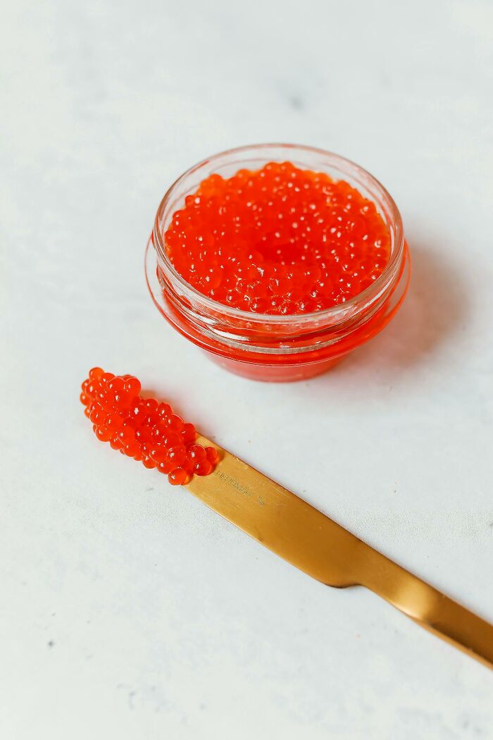 Glass jar of red caviar with a gold knife spreading caviar on a white surface, illustrating brilliantly sneaky acts of revenge.