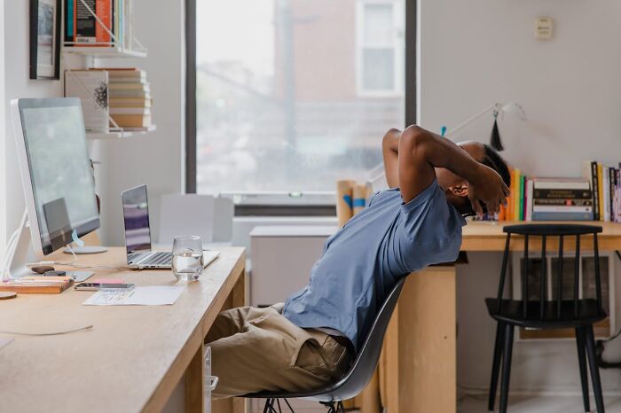 Man leaning back in chair at desk with computer and laptop, appearing stressed while working on sneaky acts of revenge.