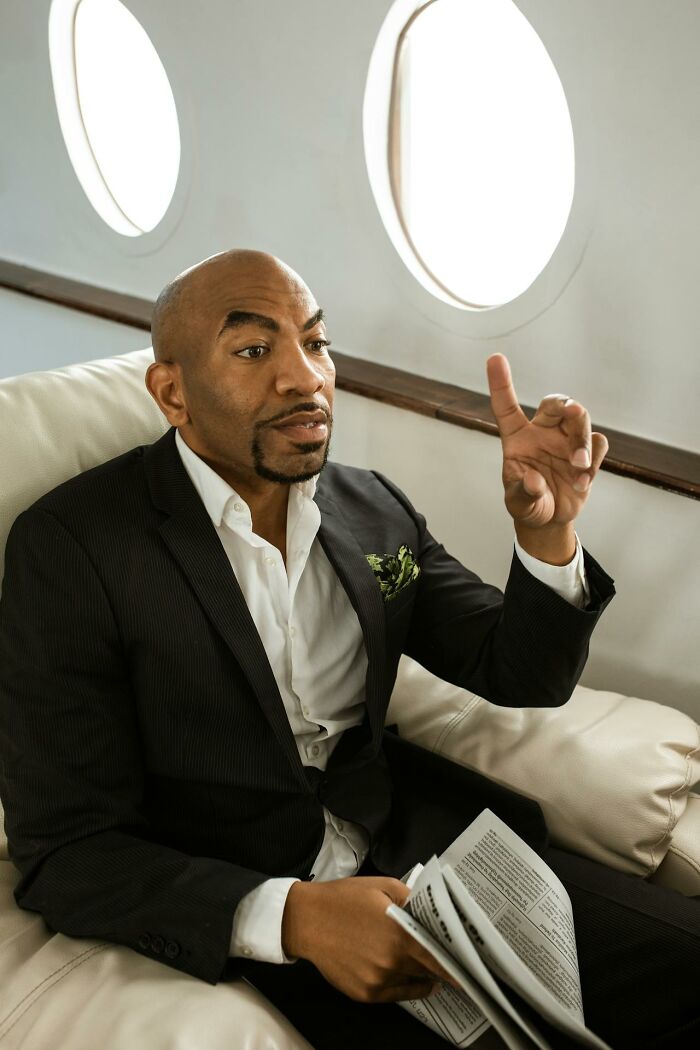 Man in a black suit displaying entitlement with a raised finger while sitting and holding a newspaper on a private jet.