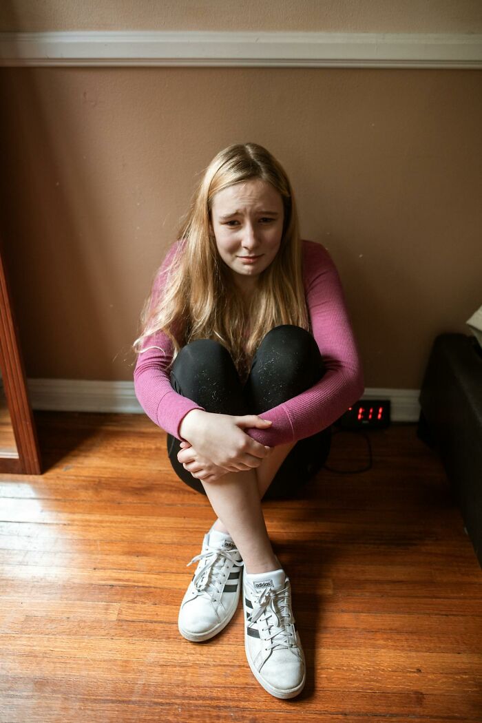 Teen girl sitting on wooden floor, hugging knees and looking upset, illustrating struggles with modern parenting trends.