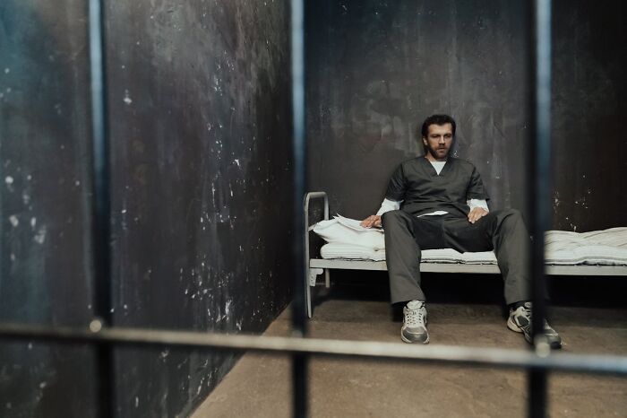 Man in casual clothes sitting on a bed in a dim room behind bars, illustrating harsh realities of being a postal worker.