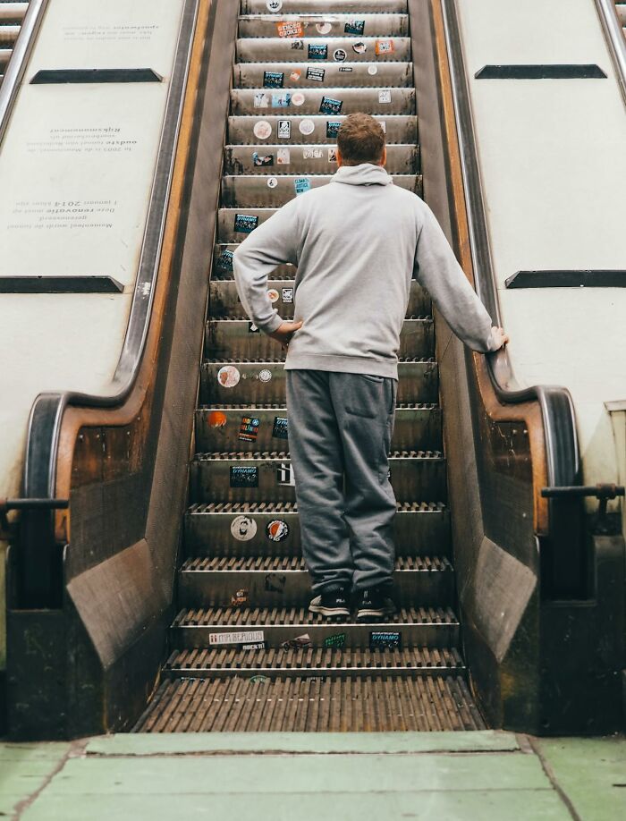 Man wearing gray hoodie and pants standing on escalator covered in stickers, reflecting crazy airport stories theme.