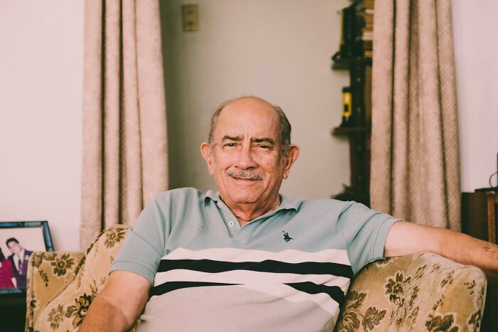 Elderly man sitting on floral couch, smiling gently, evoking unresolved mysteries people carry with them.