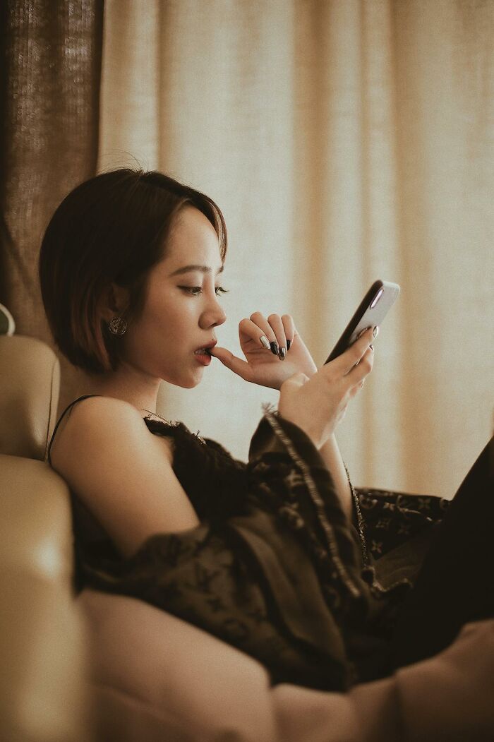 Young woman sitting on a couch, looking thoughtfully at her phone, reacting to disturbing facts she learned online.