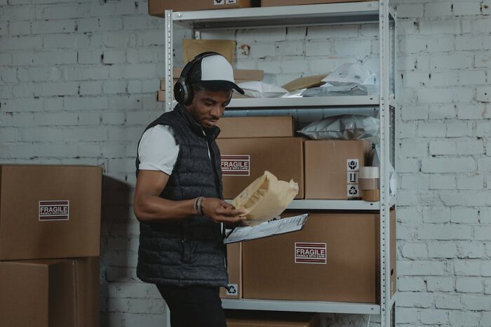 Man in a cap and vest sorting fragile packages in a storage room with lost things left unclaimed on shelves.