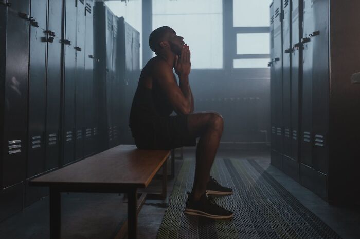 Man sitting on a bench in a dimly lit locker room, reflecting thoughtfully in a scene of sneaky acts of revenge.