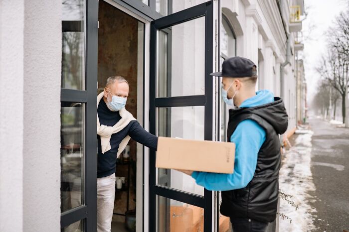 Postal worker wearing a mask delivering a package to a resident’s door on a cold day during winter season outdoors.