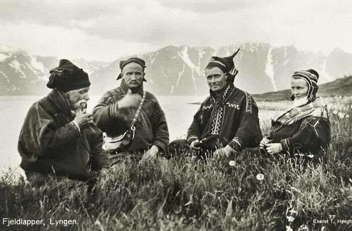Historical picture of four Fjeldlapper people in traditional clothing sitting on grass with mountains in background.