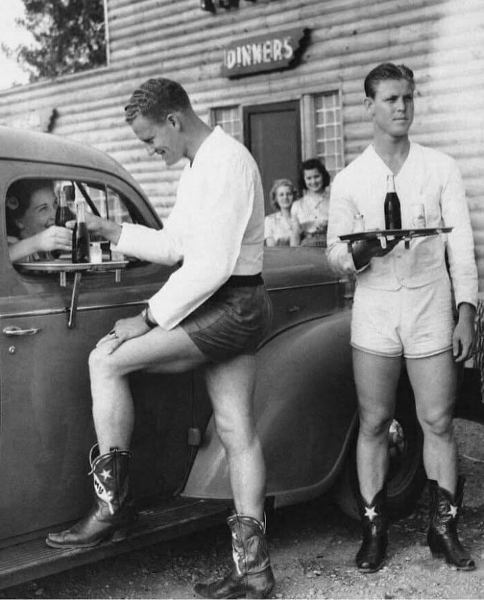 Two men in shorts and cowboy boots serving soda at a drive-in diner in a classic historical picture.