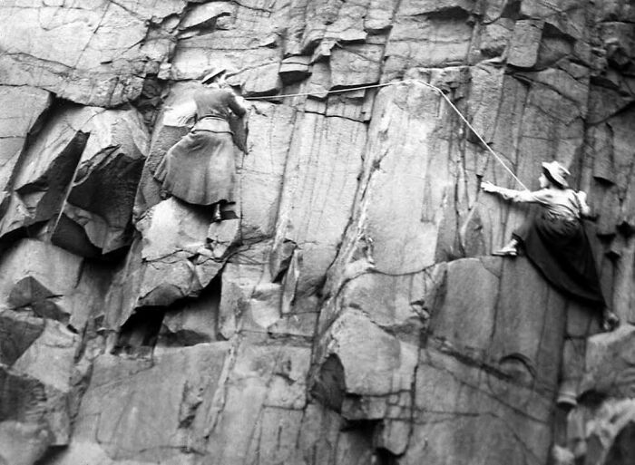 Two women in historical clothing climbing a steep rock face in a classic black and white vintage photograph.