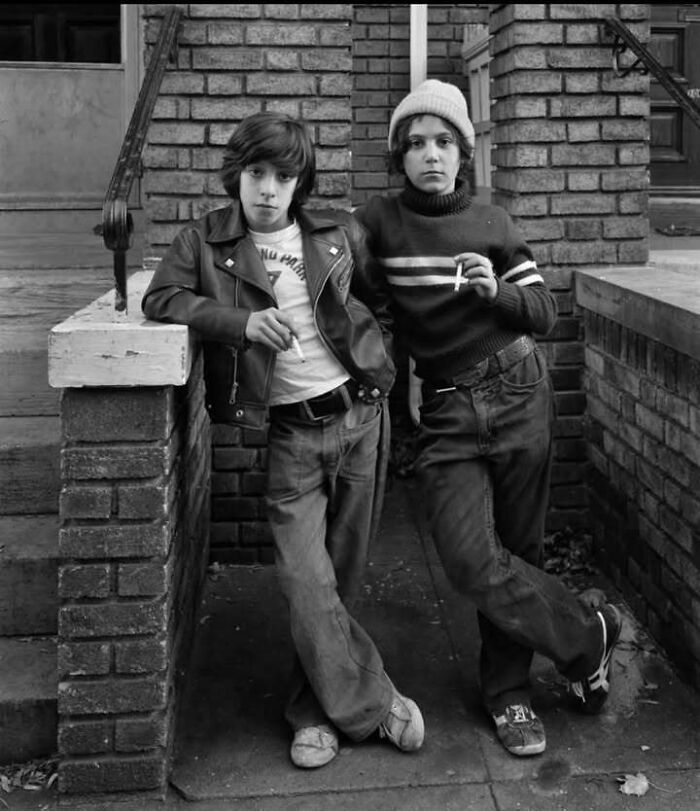 Two young people leaning against a brick wall, smoking, captured in a classic historical black and white photo.