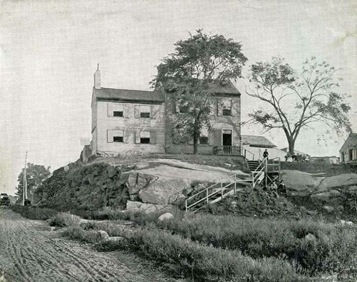 Black and white historical picture of an old farmhouse on a rocky hill with large trees and a dirt road nearby.