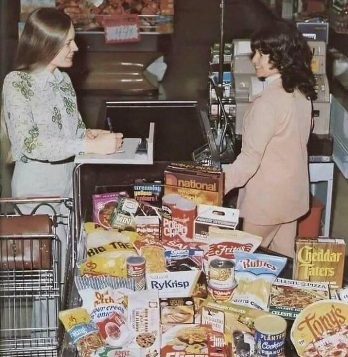 Two women smiling at a grocery checkout with a cart full of classic, iconic, and historical food products from the past.