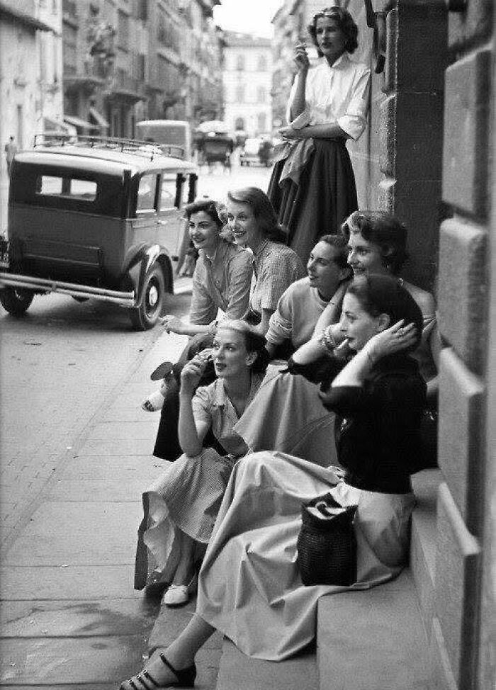 Group of women sitting and standing on a city street, vintage car in background, classic and iconic historical picture.