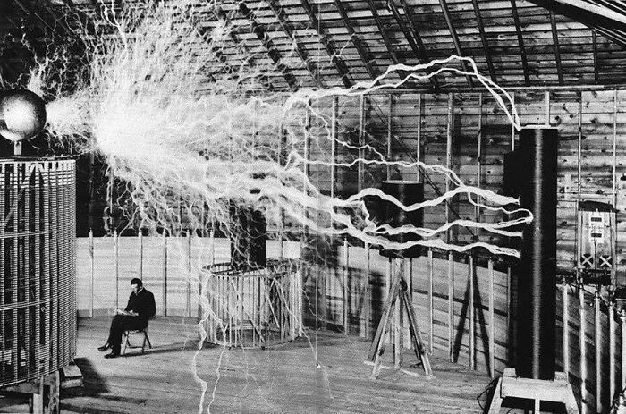 Nikola Tesla seated near his high-voltage Tesla coil emitting electrical arcs in a classic historical picture.